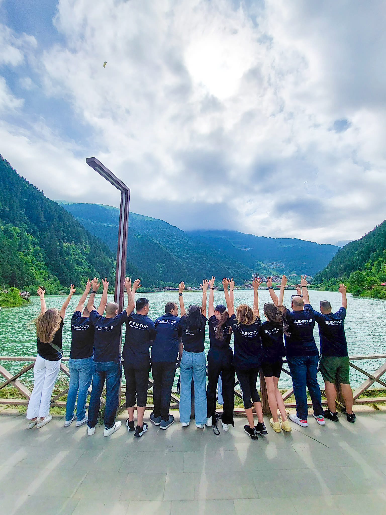 Elevatus colleagues in branded shirts raising their arms in front of a mountain lake