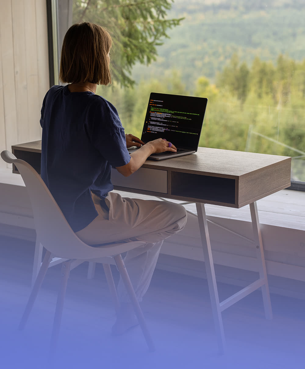 A person working remotely at a desk beside a large window overlooking trees